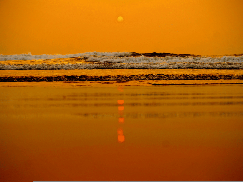 A shot from the surface of the shoreline at a beach in Goa showcasing the twilight view of the sun setting in the horizon and the waves crashing on the shoreline.