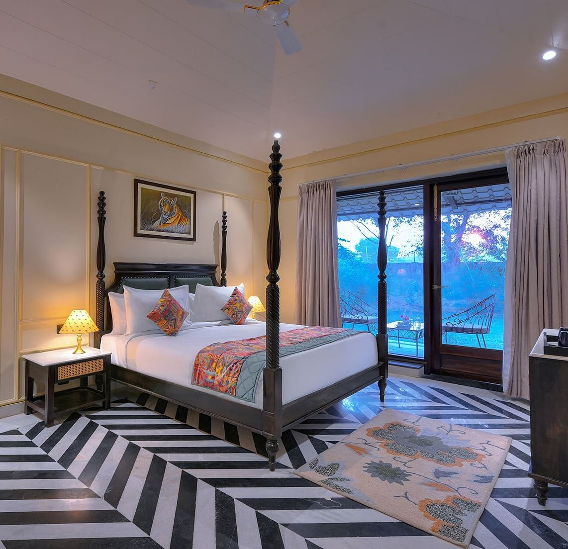 Luxury Suite Cottage featuring a four-poster bed with white linens, a colourful patterned runner and pillows, a patterned armchair, a wall-mounted TV, and sliding glass doors leading to a balcony - Singhvilas, Ranthambore