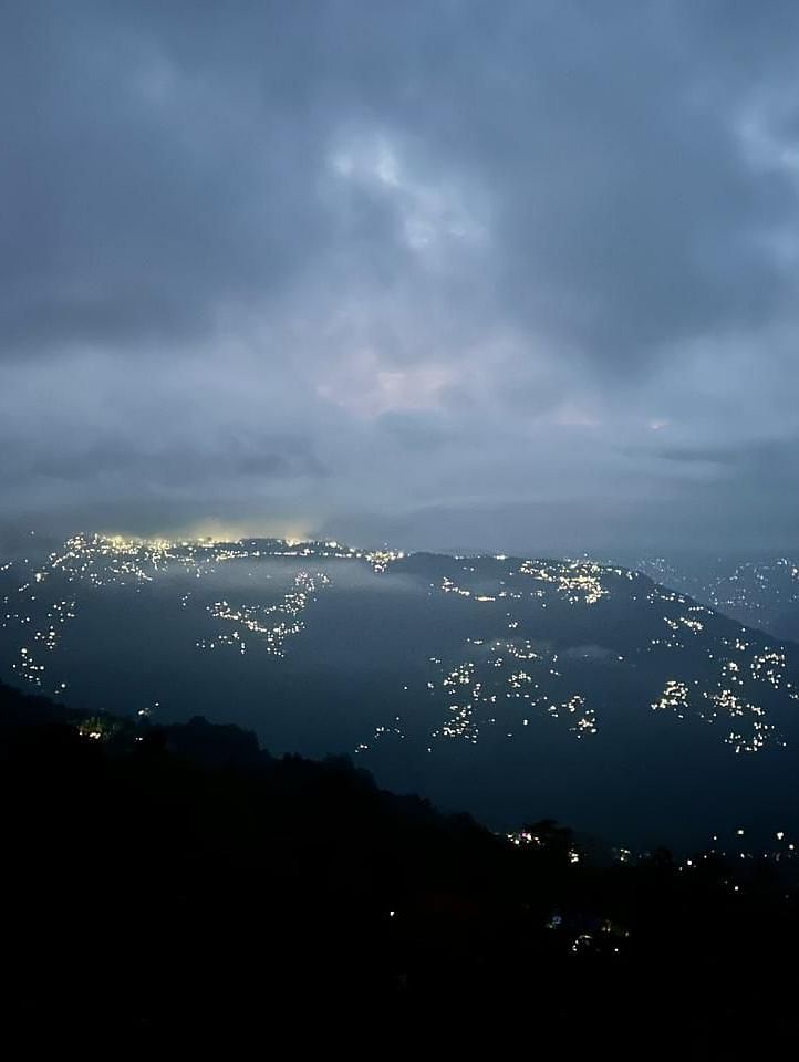  An evening view from Summit Enigma Resort & Spa shows a sprawling city twinkling with lights nestled in the hills under a dramatic cloudy sky.