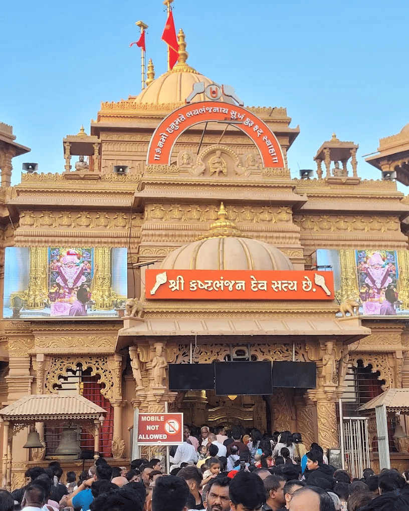 Crowds gathered at the ornate entrance of a Hindu temple with intricate carvings, domes, and digital displays of deities.