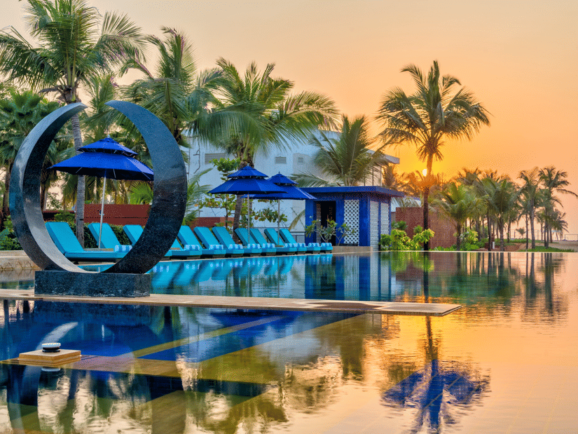  A vibrant sunset view of the swimming pool at Azaya Beach Resort Goa, featuring a large modern circular sculpture, blue lounge chairs, and palm trees reflected in the water. 