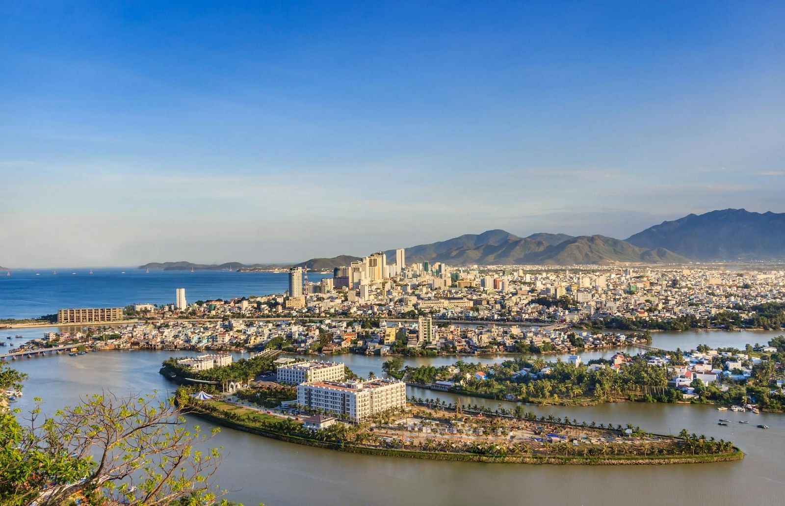 A panoramic view of a city nestled between a body of water and mountains under a clear sky.