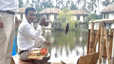 A hotel staff member serving food on a tray at a waterside outdoor dining area surrounded by trees and a pond at Vedic Village Spa Resort.