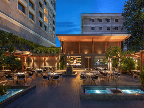 An outdoor dining area with tables, chairs, and plants, illuminated by warm lighting along with swimming pool. 
