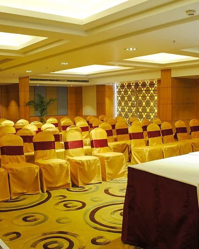 The image shows a conference room with rows of chairs facing a table with water bottles and flowers - Lords Eco Inn Navsari.