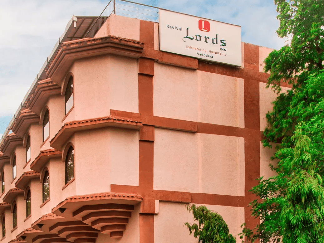 The facade of Lords Inn Vadodara, the best hotel near Vadodara Airport - featuring a red and pink toned building with the hotel signboard and lush greenery under partly clouded sky.