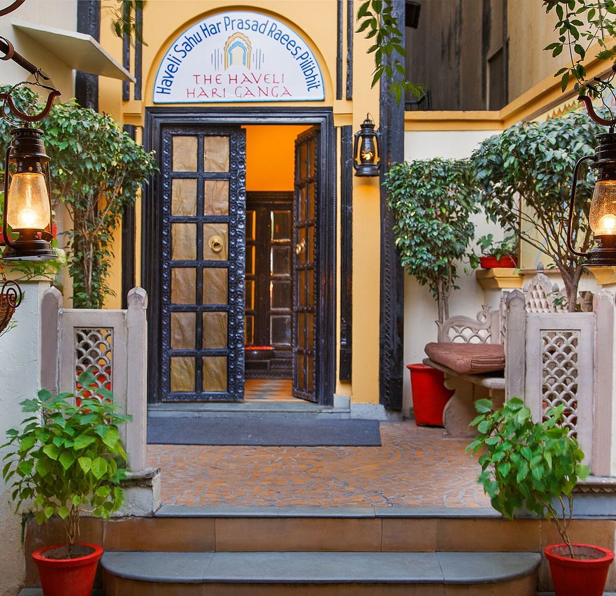 A wooden-furnished door at The Haveli Hari Ganga in Haridwar, adorned with plants, creates a warm and inviting entrance that enhances the beauty of the setting, welcoming guests with a touch of nature and charm.
