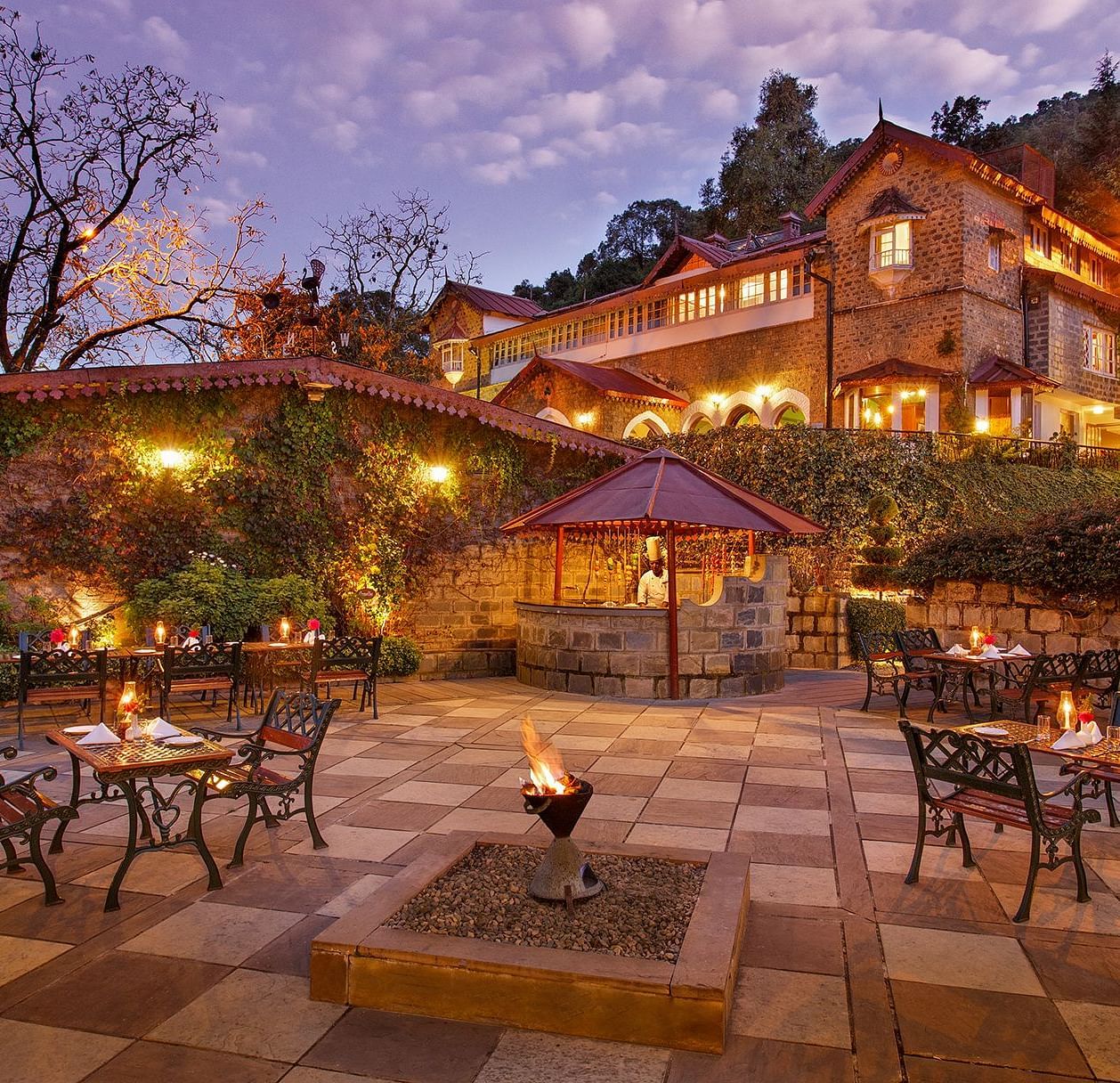 A beautiful outdoor dining area at the naini retreat, nainital. There are tables and chairs set up on a stone patio, with a fireplace in the center. The building is lit up at dusk, creating a warm atmosphere. 