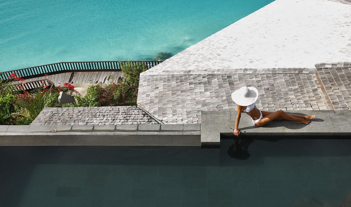 An overhead view of a woman in a wide-brimmed hat lounging by a dark infinity pool overlooking a vibrant turquoise ocean.