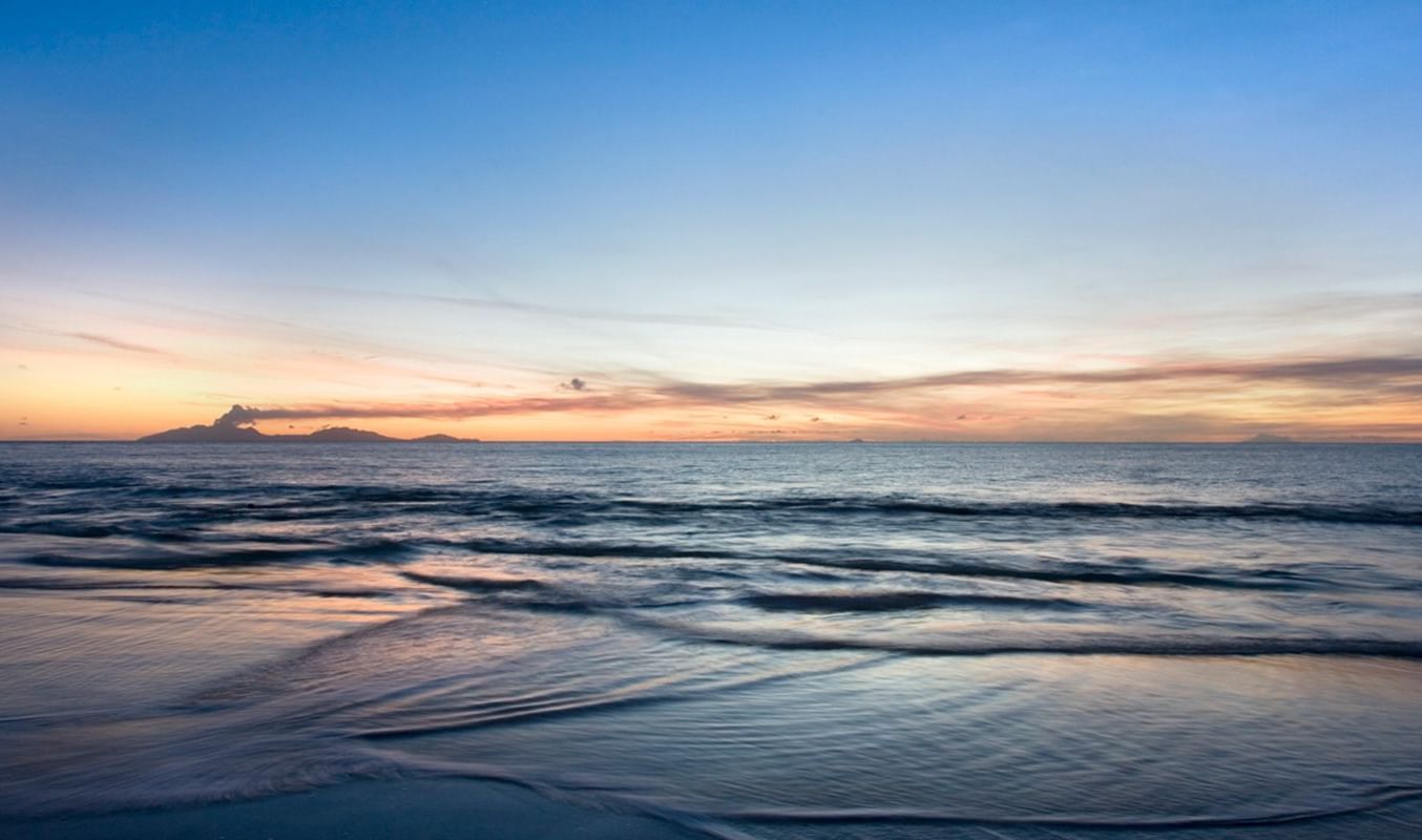 Peaceful Caribbean shoreline at sunset with soft waves, golden hues on the horizon, and a tranquil beach atmosphere.