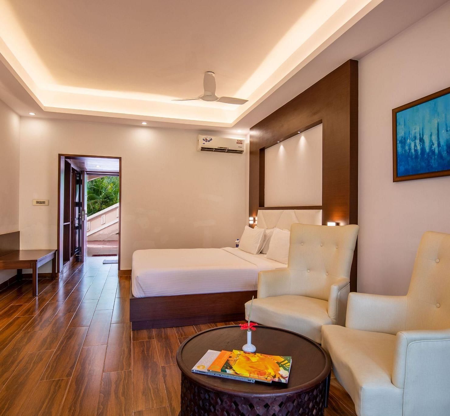 A contemporary hotel bedroom at Avana Resort with a large bed, two armchairs, and a round coffee table, featuring wooden floors and a balcony. 