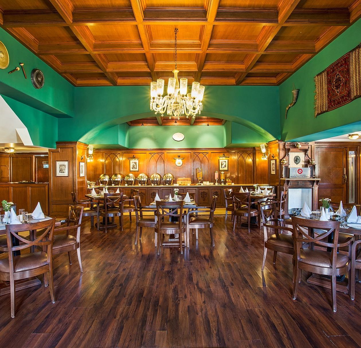 An image showing the interior of restaurant at The naini retreat, Nainital. featuring wooden decor, flooring, and a wooden bar. A large chandelier hangs from the ceiling, providing ambient lighting.