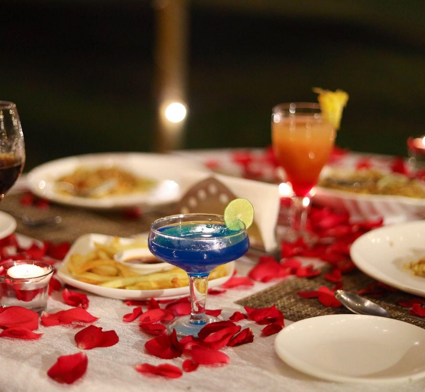 A close-up of a romantic outdoor dining table set with a blue cocktail, scattered red rose petals, and candlelight at Avana Resort.