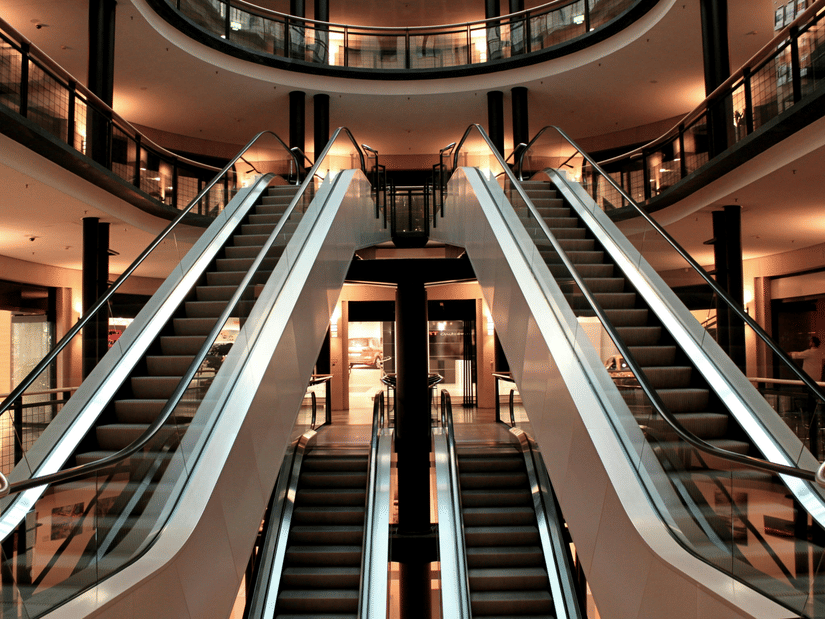 A view of the interior of a multi-level building, likely a shopping mall, showing a pair of escalators going up and another pair going down, with balconies on the upper floors and a circular open space above.