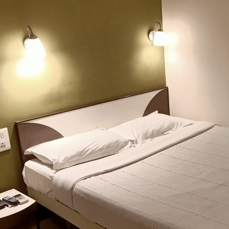 A Standard AC Room at Hotel Pandian, featuring a neatly-made bed with white sheets, 2 pillows, 2 wall lamps, and a nightstand.