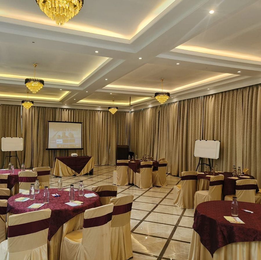 alt-text A banquet hall arranged for a corporate event with beige chairs, burgundy tablecloths, large windows letting in natural light, and chandeliers adding a luxurious touch to the setting.