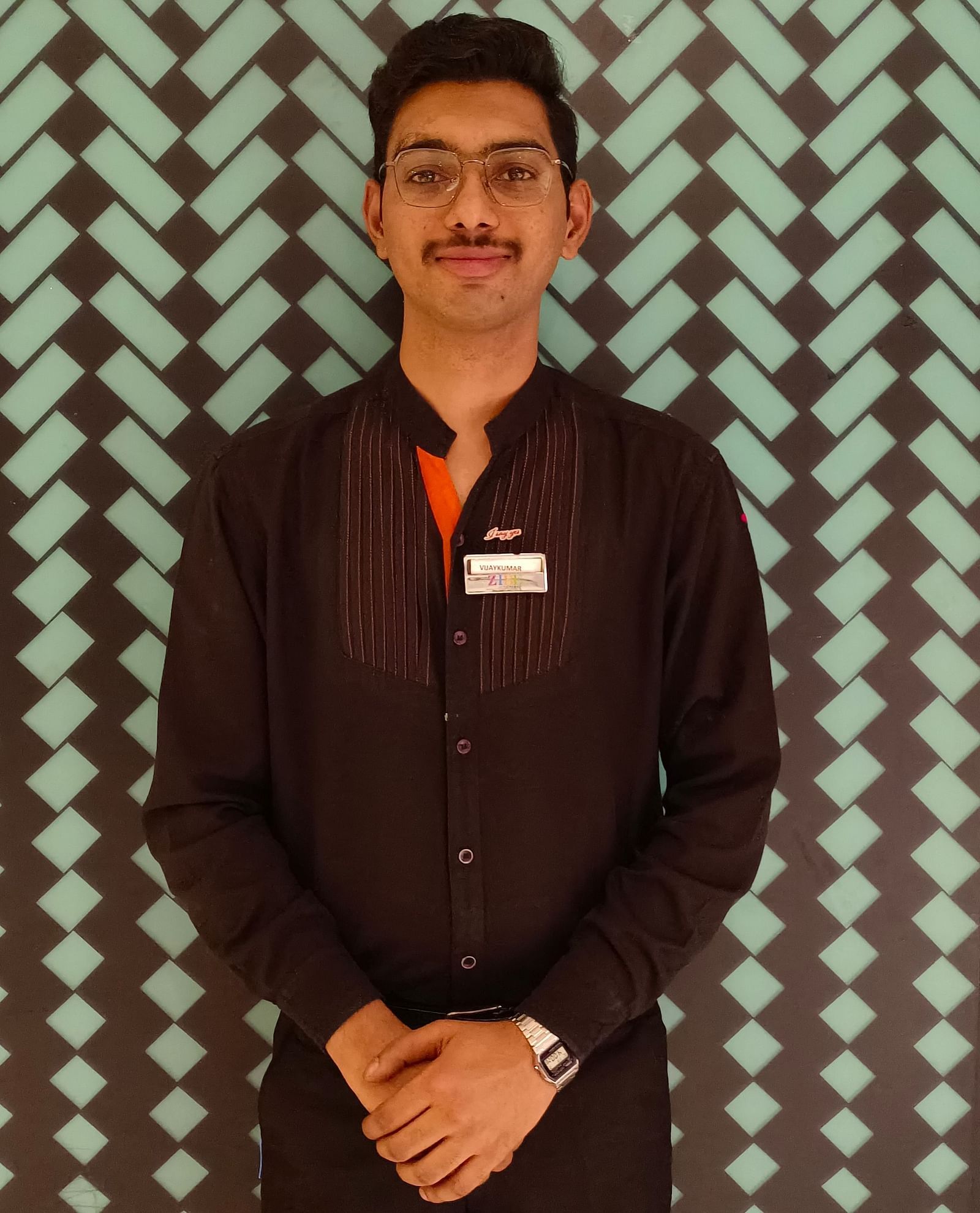 Mr Vijay stands with poise against a backdrop featuring a repeating pattern