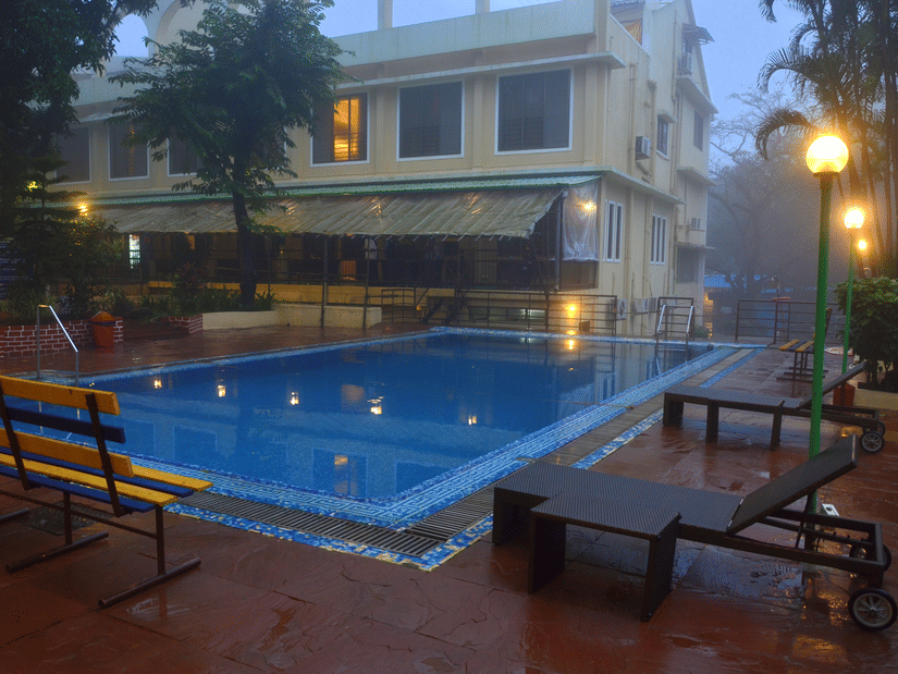 A swimming pool reflects the soft glow of nearby lights, with lounge chairs placed along the wet deck and a building partially veiled in mist in the background | Zara's Resort, Khandala