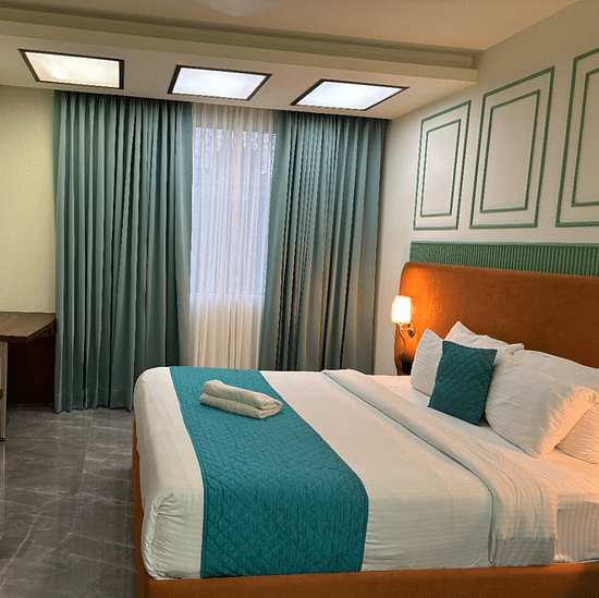 An interior view of a hotel room featuring a neatly made bed with pillows , a television and a window with curtains -  Asian Suites IFFCO Chowk Metro Station, Gurgaon.