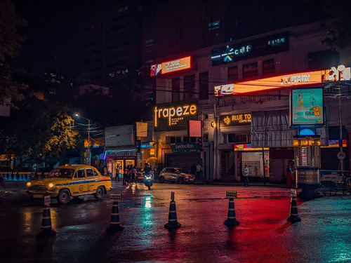 A vibrant Kolkata street at night with illuminated storefronts, city traffic, and the iconic yellow taxi.