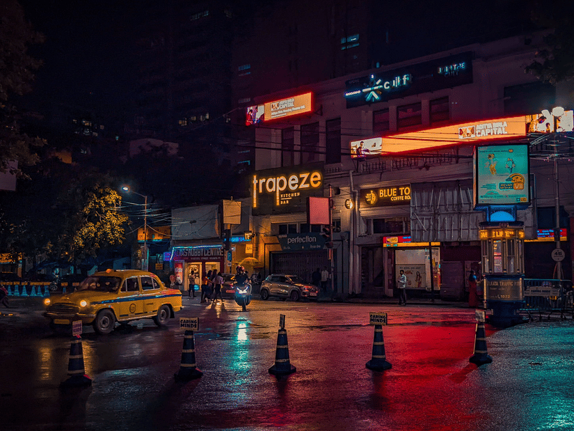 A vibrant Kolkata street at night with illuminated storefronts, city traffic, and the iconic yellow taxi.
