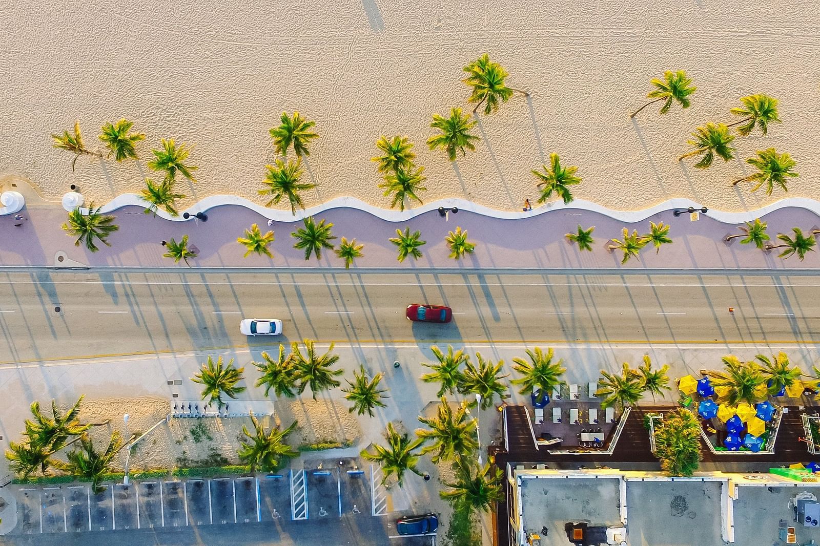 A top view of a road next to the beach with vehicles, buildings and trees in view.