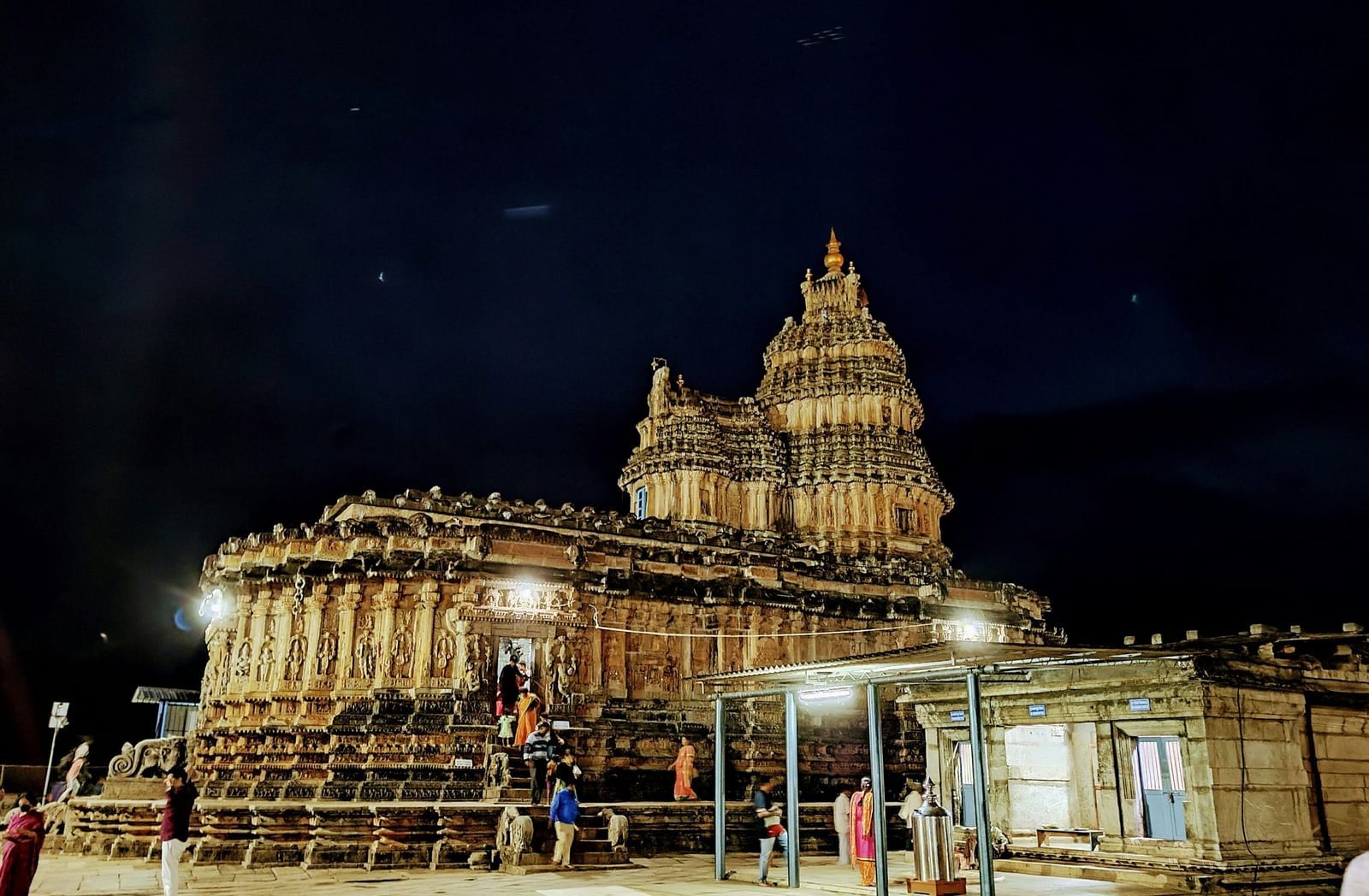  A nighttime shot of an ancient stone temple with a tiered tower, lit up against a dark sky, with people gathered in the foreground.