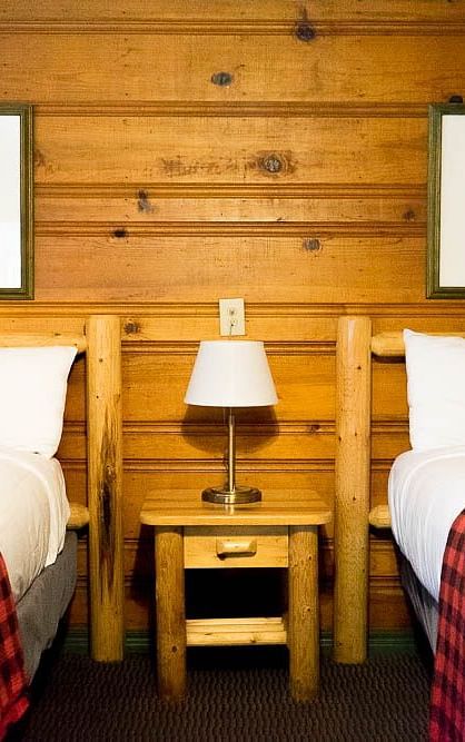 A wooden cabin with twin beds and a side table with a night lamp placed between them at Shaver Lake Village Hotel.