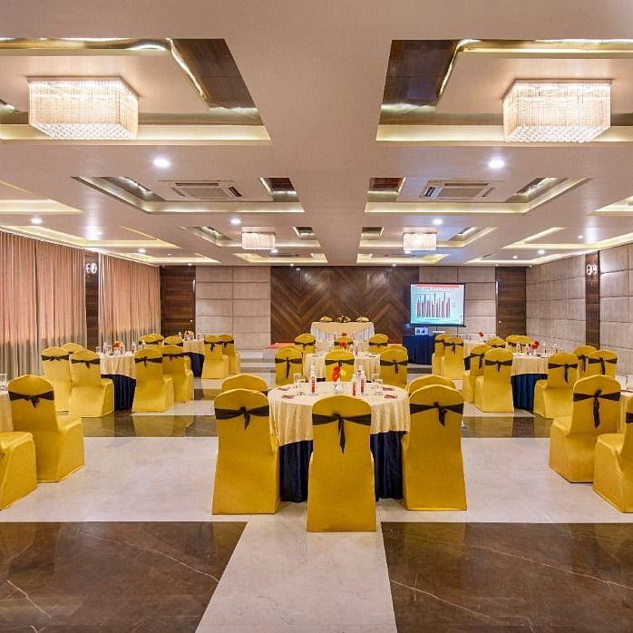 A spacious event hall at Lords Inn Porbandar, is prepared with round tables and chairs with yellow sashes, ready for guests.