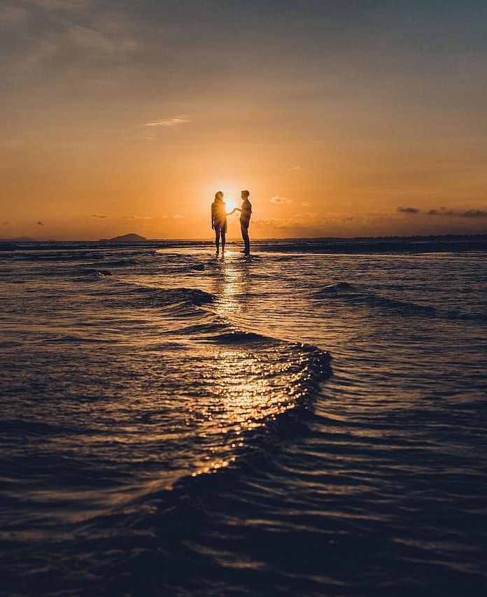 A couple standing on the shoreline during sunset and their reflection visible in the ocean