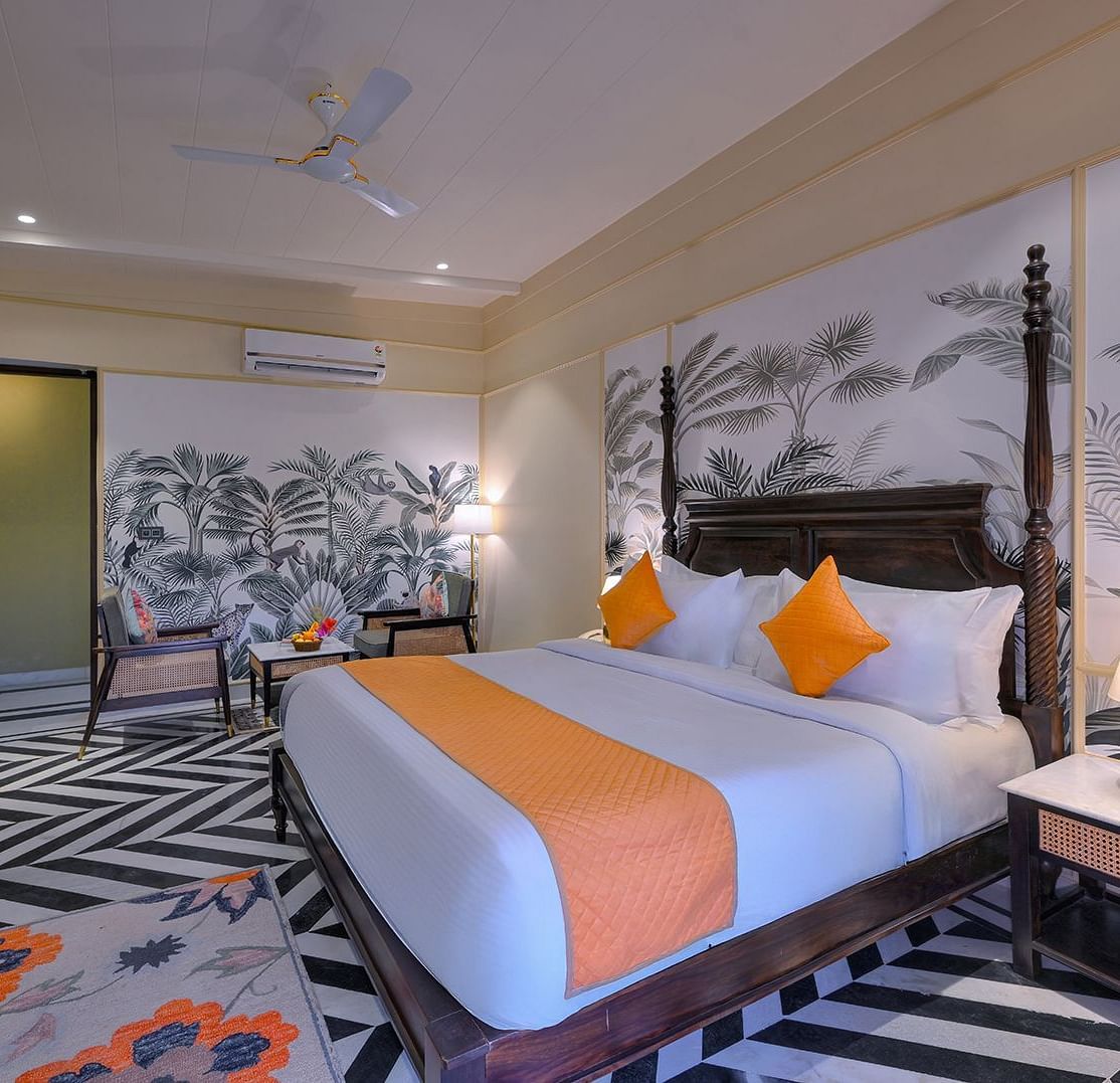 Superior Cottage featuring a four-poster bed with orange accents, chevron-patterned flooring, tropical wallpaper, a seating area, and a nightstand with a gold lamp - Singhvilas, Ranthambore