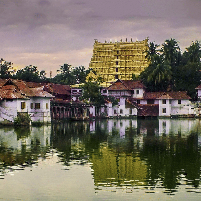 Sree Padmanabha Swamy Temple and a row of houses reflected in a calm lake during cloudy evening light, with trees lining the waterfront.