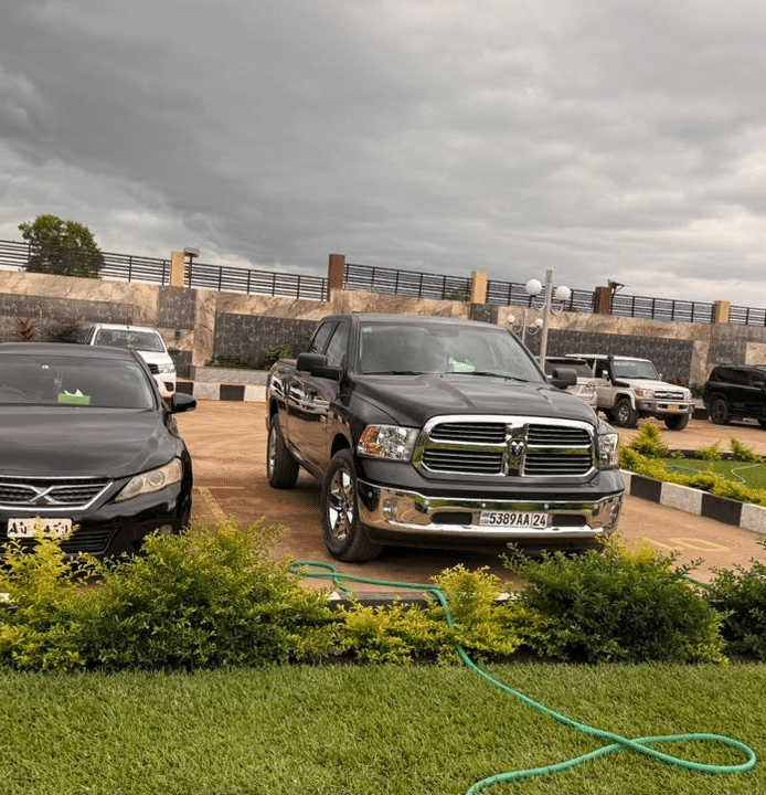 A wide shot of a modern building's exterior with two parked black SUVs, a lawn area, and a cloudy sky at Luffu Club.