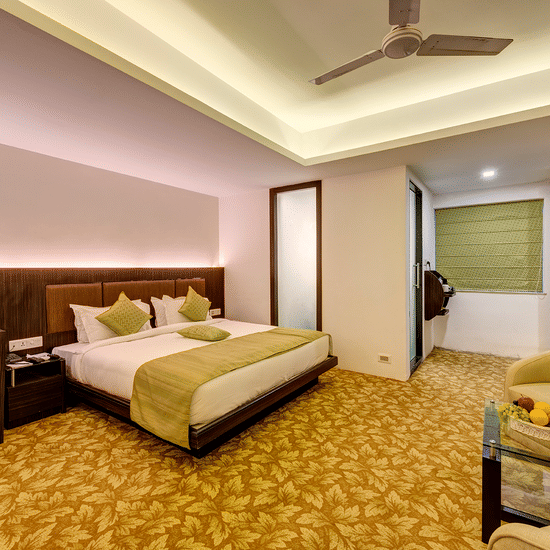 Premium room with king bed, green accents, work desk with monitor, red flower vase, armchairs with fruit table, patterned carpet, and warm lighting at Viceroy Boutique Hotel, Kolkata.