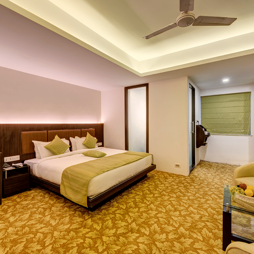 Premium room with king bed, green accents, work desk with monitor, red flower vase, armchairs with fruit table, patterned carpet, and warm lighting at Viceroy Boutique Hotel, Kolkata.
