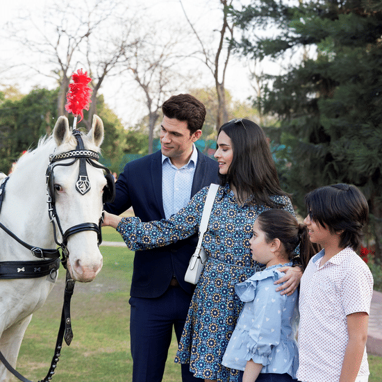 A family standing beside a horse with a red plume, petting it in a park with trees and a paved path nearby, at Heritage Village Resorts & Spa, Manesar.