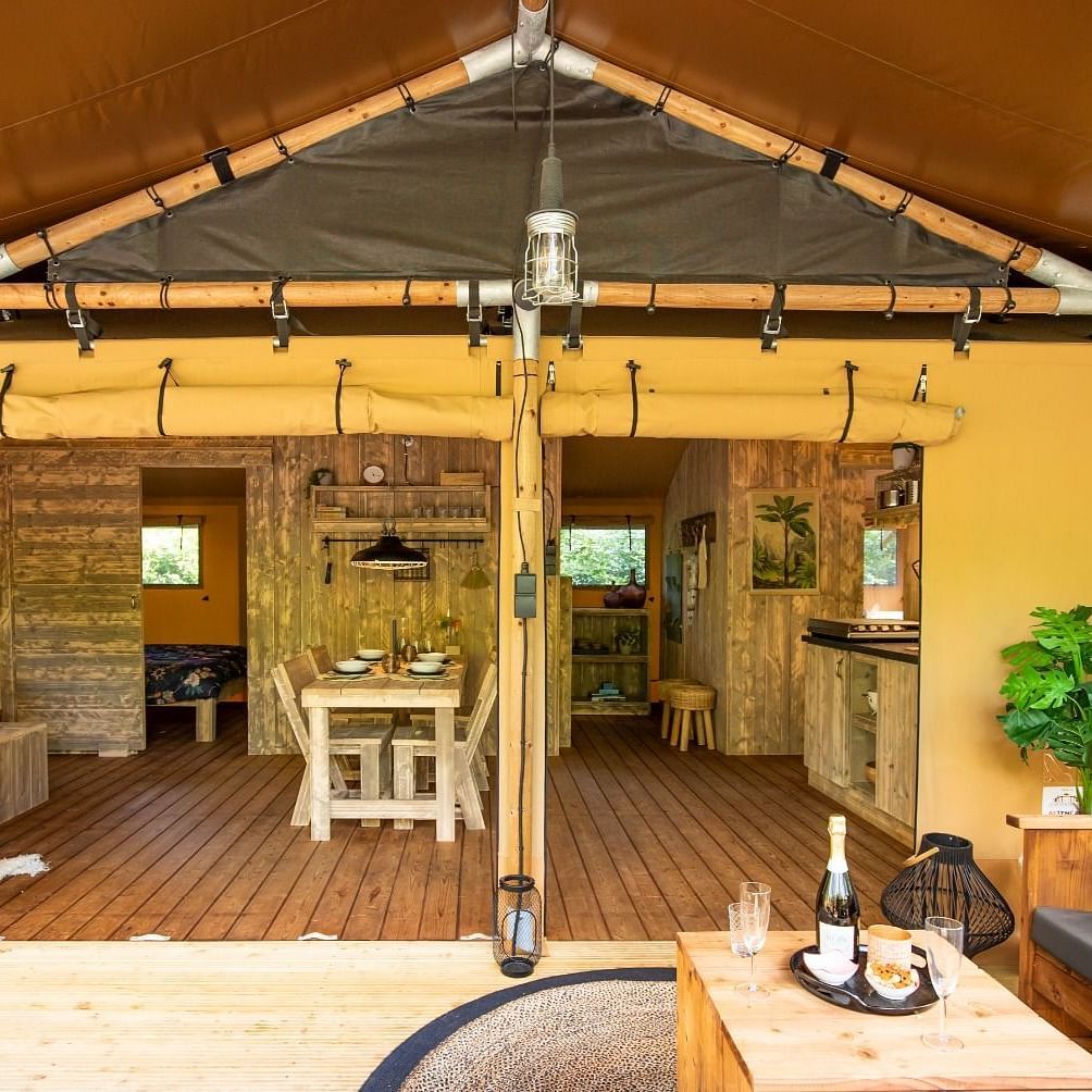 A glimpse inside a tent reveals a cozy interior with wooden flooring, and a small dining table. The tent features a canvas roof and walls, suggesting a comfortable glamping experience.