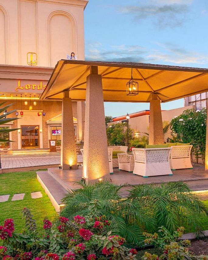 An inviting outdoor area at dusk with a covered seating space supported by pillars, surrounded by well-maintained green lawns, colourful flowering plants, and subtle lighting illuminating the building in the background at Lords Inn Motikhavdi, Jamnagar.