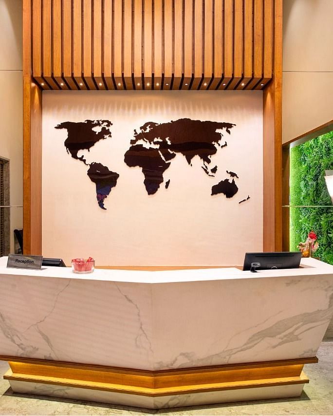 A contemporary reception area with a sleek white and gold front desk, a large dark world map on the wall, vertical wooden slats above, and illuminated green moss panels on the side walls, creating a modern and welcoming atmosphere - Lords Inn Motikhavdi, J