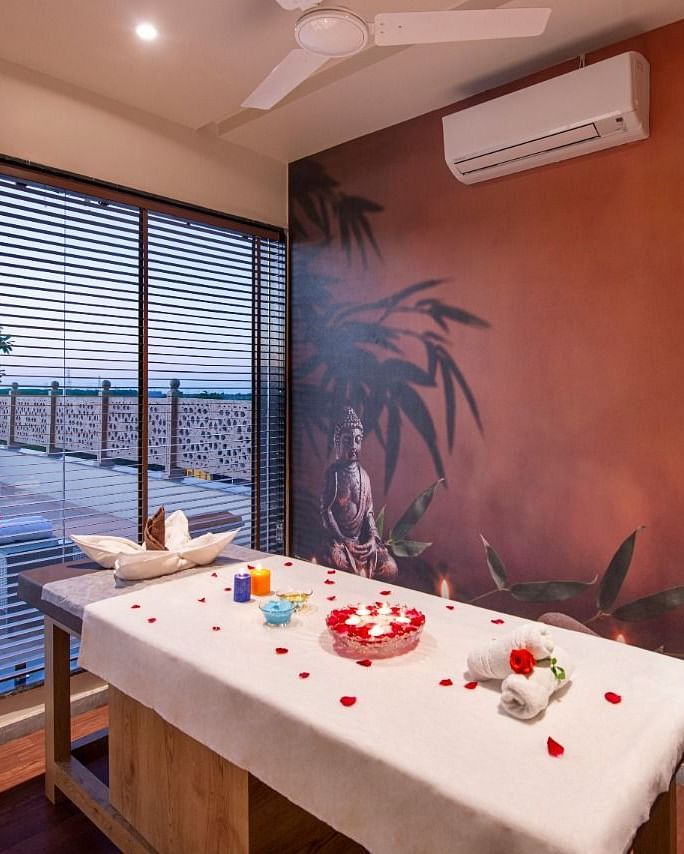 A serene spa treatment room featuring a massage table draped with a white sheet and adorned with red rose petals and decorative items, set against a warm-toned wall with a subtle mural and with natural light coming through large windows overlooking an outd