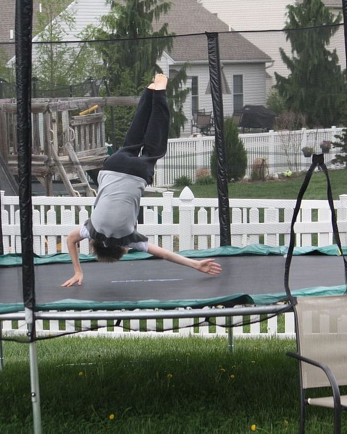 A kid jumping on a trampoline surrounded with standalone buildings and blue fencing.
