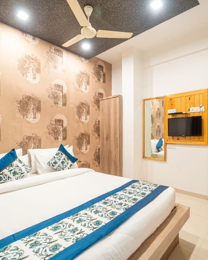 A room featuring a bed with blue and white linens, a patterned wallpaper, a ceiling fan, a television, and a wooden closet. 