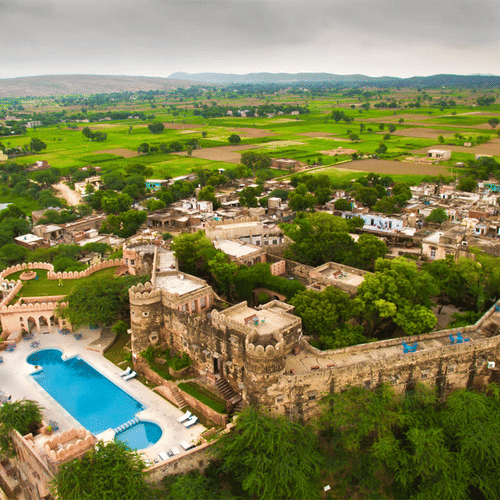 Aerial drone shot of Hill Fort Kesroli 14th Century Alwar where swimming pool and multiple structures are surrounded by lush green landscape and cloudy blue sky in the background.