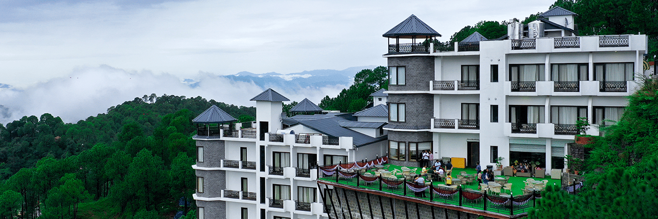 Panoramic view of Rosetum Hotels Kasauli perched on a forested hillside with misty mountains and an open terrace