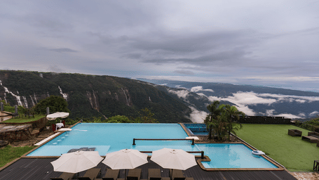 Infinity pool with white umbrellas overlooking cliffs and waterfalls under a cloudy sky, with green lawn and lounge chairs on the right.