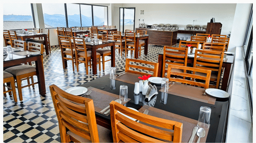 Seating arrangement inside the in-house restaurant serving the best food in Vagamon, with floor to ceiling windows in the distance having valley-views at Abad Silvermist, Vagamon