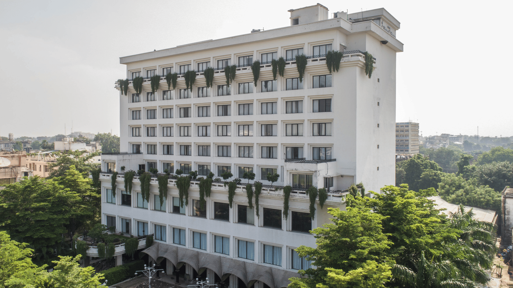 A multi-storey white building with rows of windows, green hanging plants on the facade, and trees in the foreground under a bright sky.