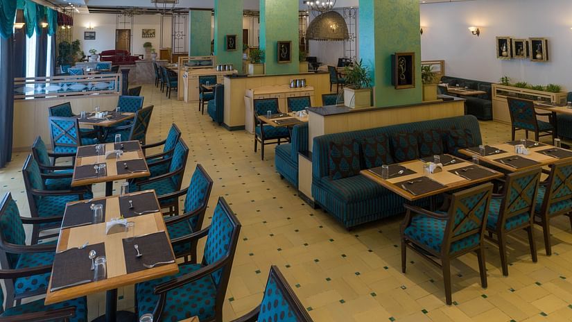 A restaurant interior with wooden tables, blue upholstered chairs, table settings with cutlery and napkins, green pillars, and framed wall decorations.