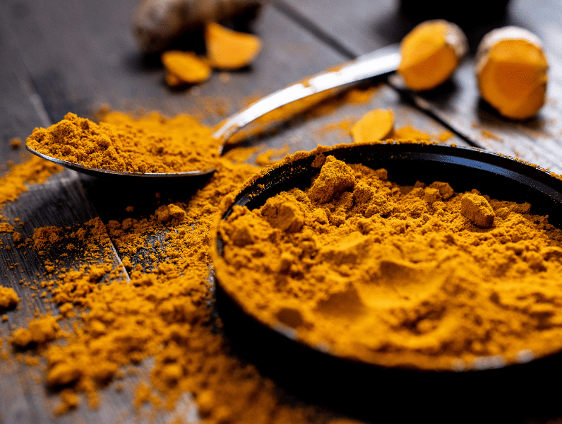 A close-up of vibrant turmeric powder in a bowl and spoon, with fresh turmeric root pieces scattered on a dark wooden surface.