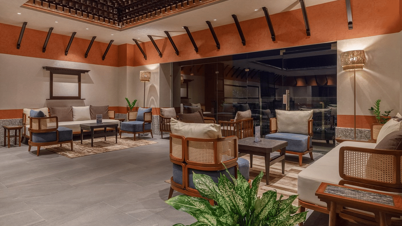 Lobby lounge with cane-backed armchairs, cushioned sofas, potted plants and a carved timber ceiling with sloping struts at Ila Resort, Maulakali.